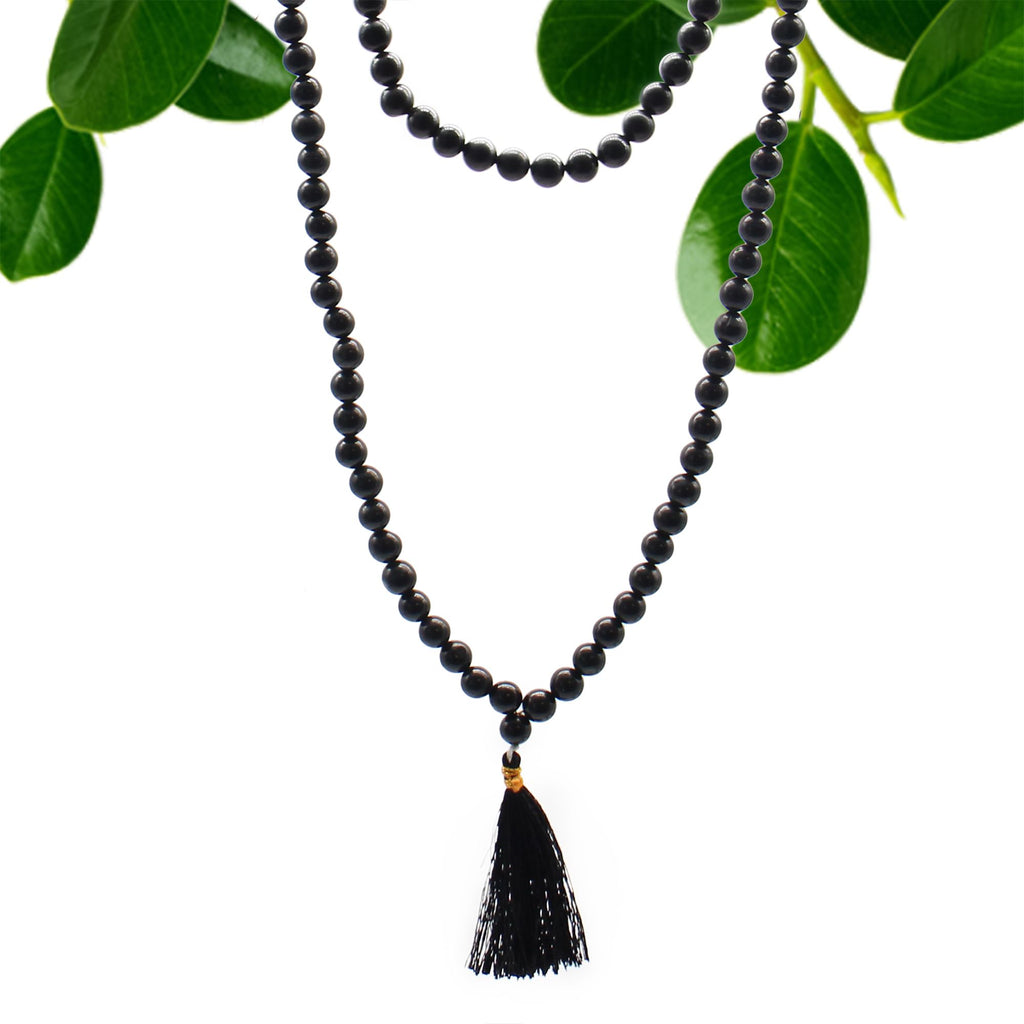 Collier mala 108 perles en pierre naturelle agathe noire avec pompon noire, suspendu avec feuilles vertes en arriere plan, bijou spirituel pour meditation chakra et bien etre