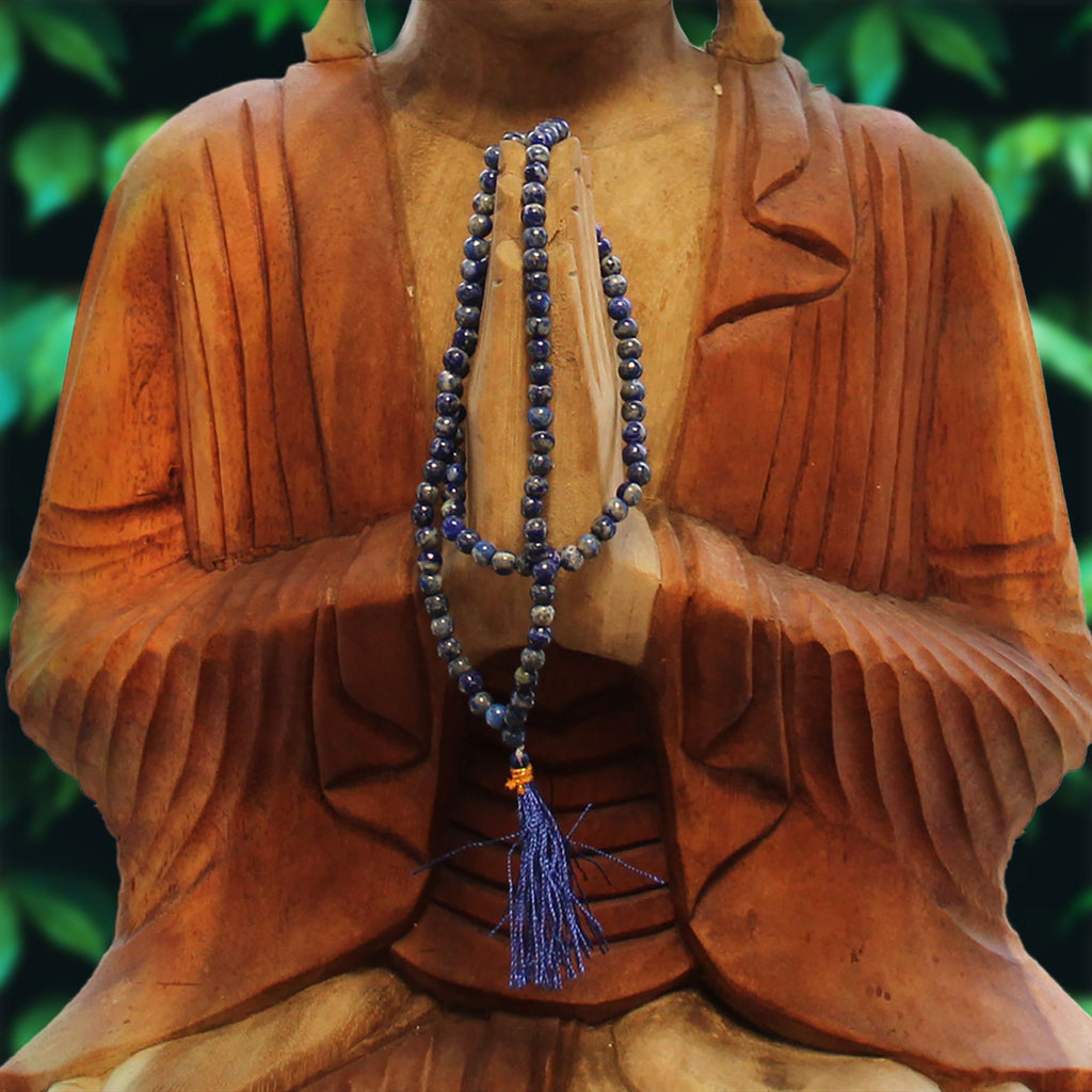 Collier mala 108 perles en pierre naturelle lapis lazuli avec pompon bleu, suspendu sur statute bouddha avec feuilles vertes en arriere plan, bijou spirituel pour meditation chakra et bien etre