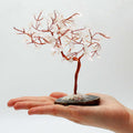 Arbre en cristal de roche avec base en agate noire posé dans une main, branches fines en métal et pierres transparentes
