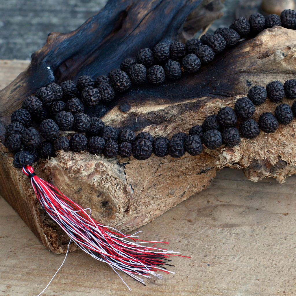 Mala en perles de rudraksha foncé posé sur une surface en bois brut avec pompon rouge et blanc, ambiance naturelle et authentique.
