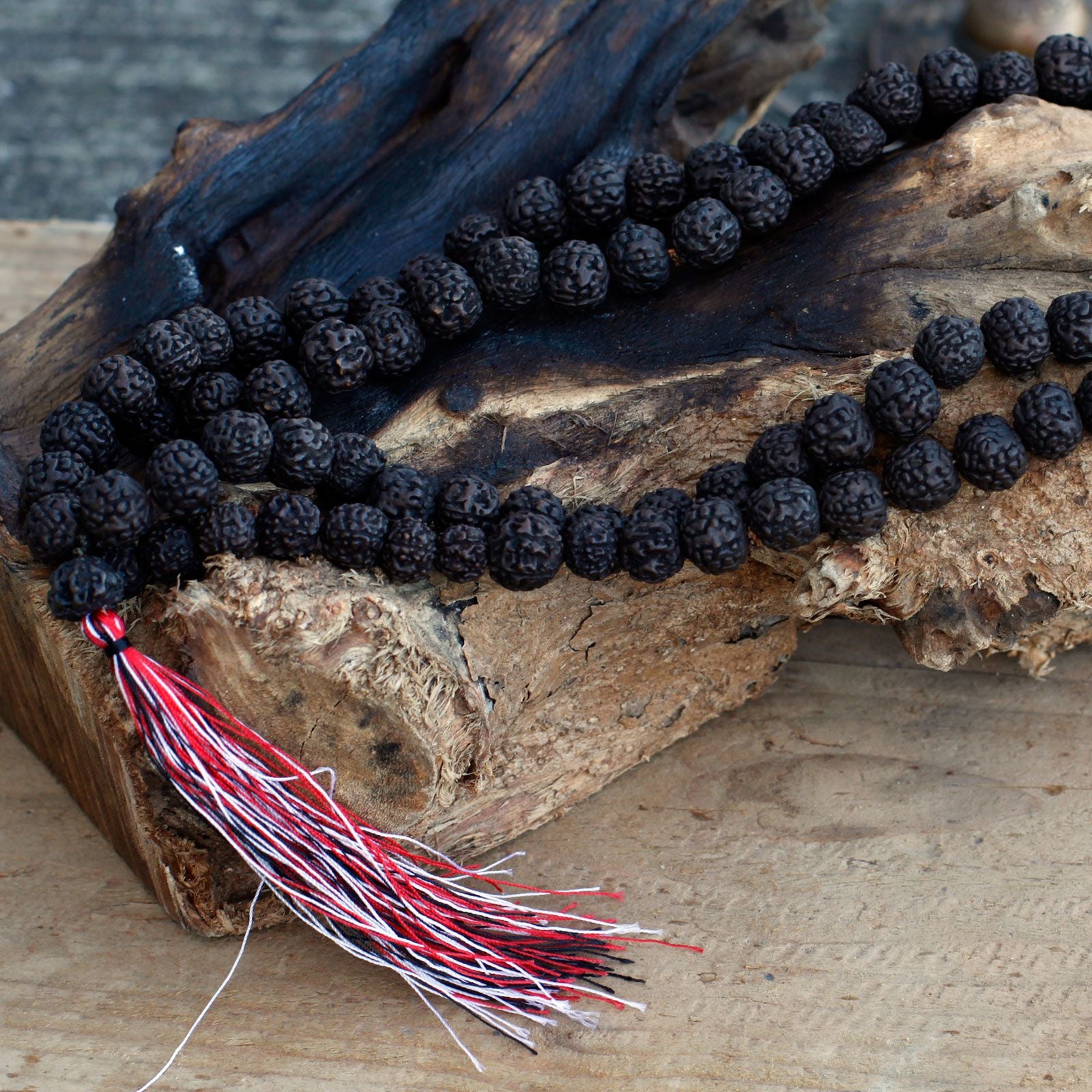 Mala en perles de rudraksha foncé posé sur une surface en bois brut avec pompon rouge et blanc, ambiance naturelle et authentique.